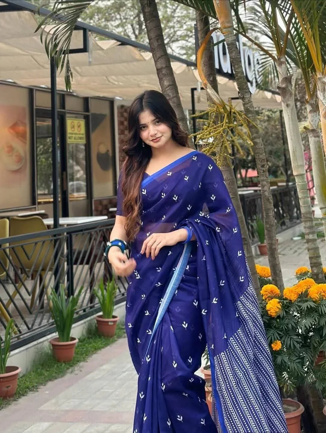 Woman wearing a blue saree with white patterns, standing outdoors by potted plants