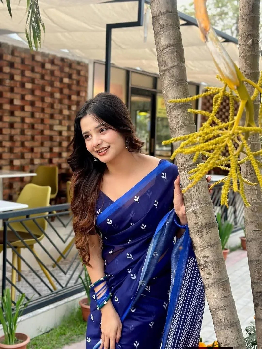 Woman in blue saree with white patterns posing outdoors near a tree, smiling in garden cafe
