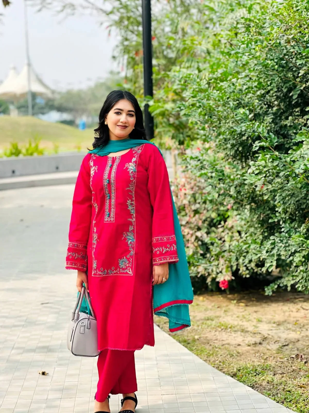 Woman in pink embroidered salwar kameez with teal dupatta, posing outdoors with greenery