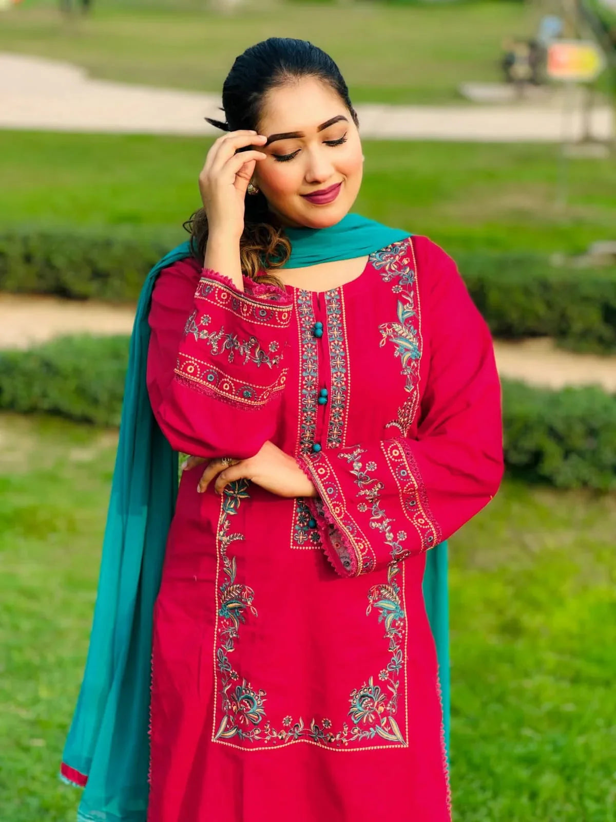 Woman in red embroidered kurta with teal dupatta, standing outdoors on green grass