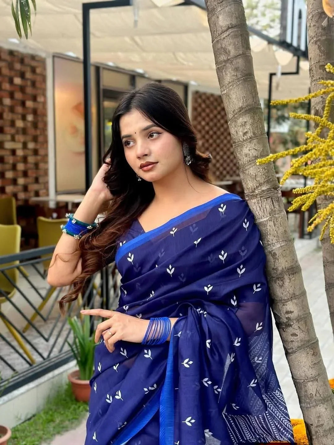Woman in blue printed saree with matching bangles posing outdoors near a café and palm tree
