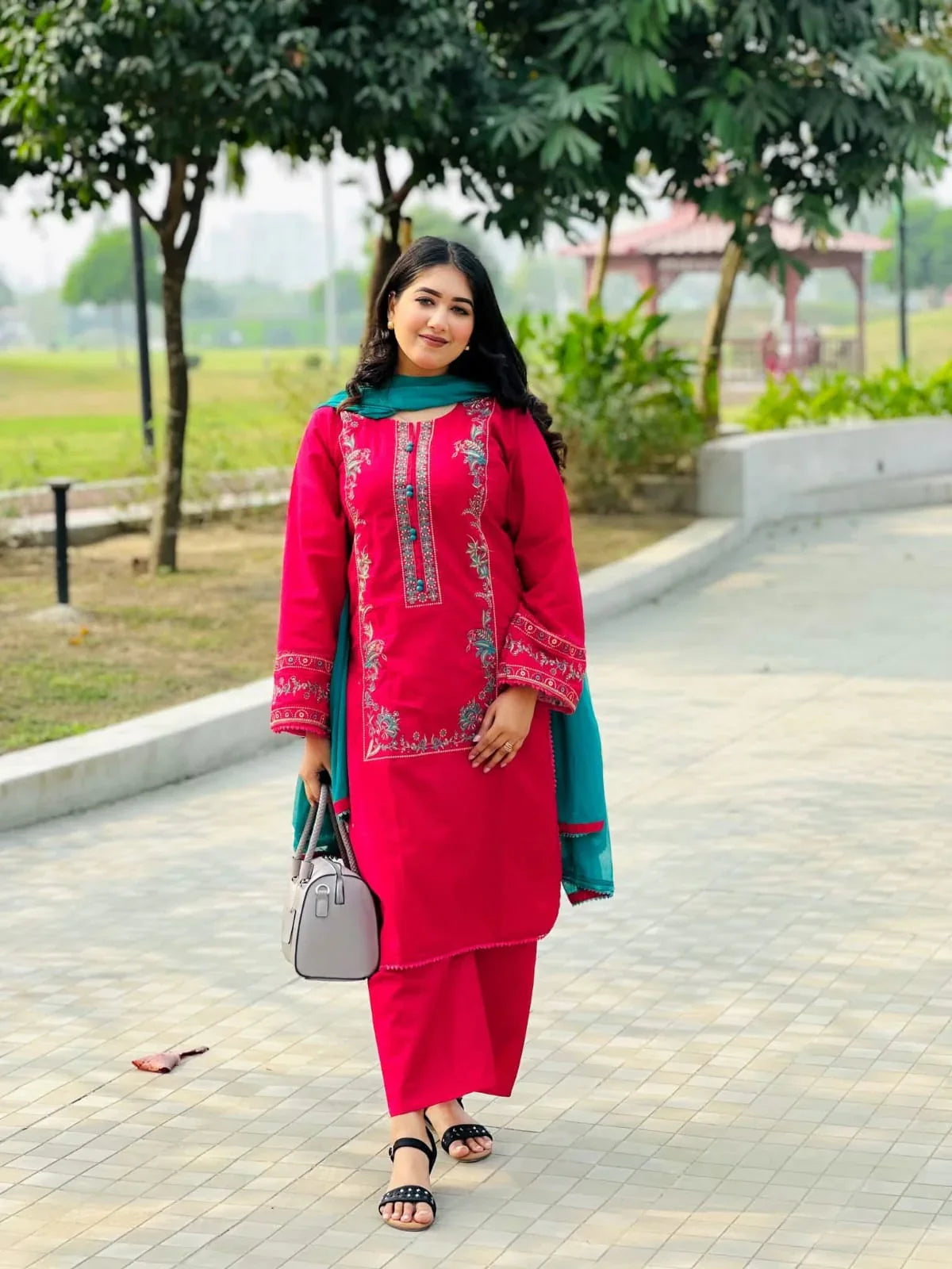 Woman in a red embroidered kurta set with teal dupatta, posing outdoors in a park.